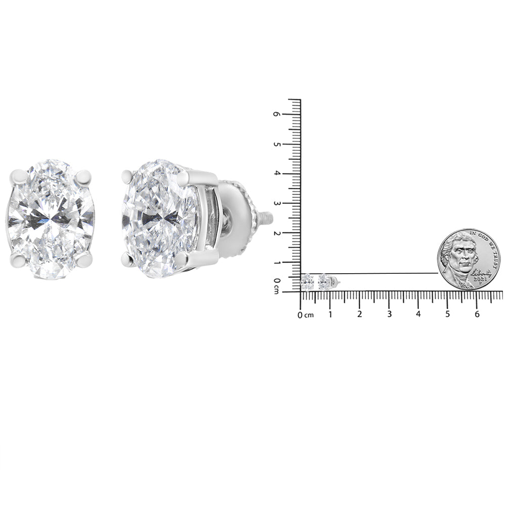 Pair of white metal stud earrings with oval-shaped white gemstones in a 4-prong setting shown at a front and side angle, next to a ruler and coin for size reference.