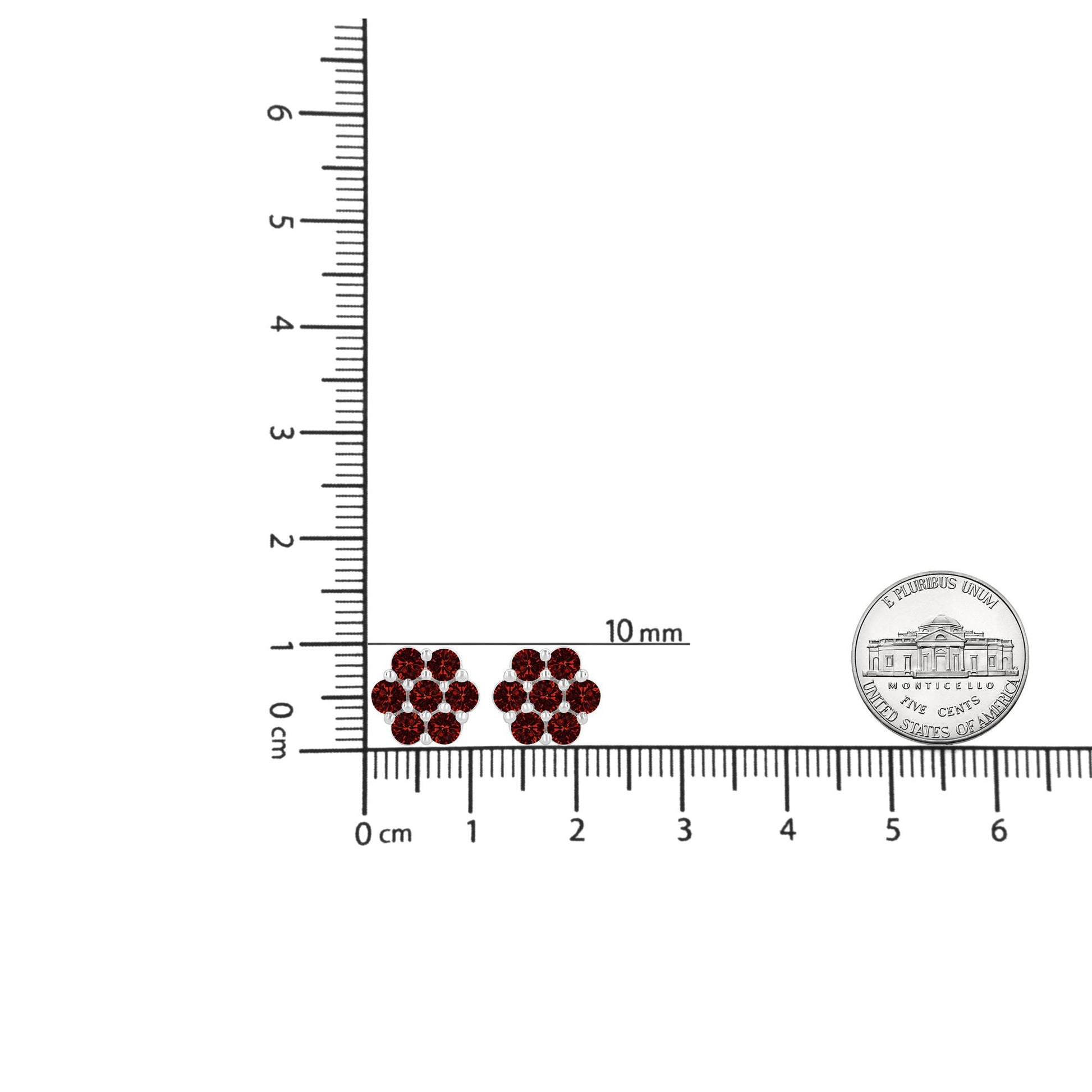 Pair of white metal earrings with round red gemstones in prong settings, displayed on a ruler and a coin for size reference, front view.