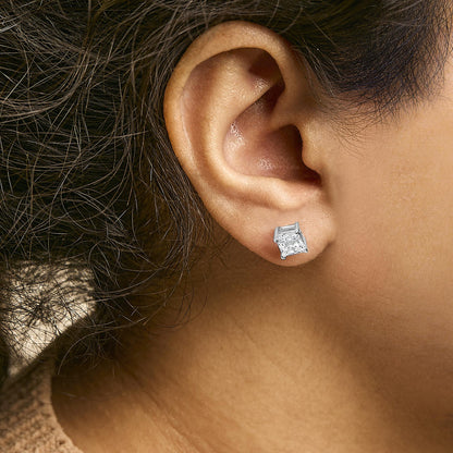 Close-up side view of a white metal earring with a princess-shaped white gemstone in a four-prong setting, worn on a model.