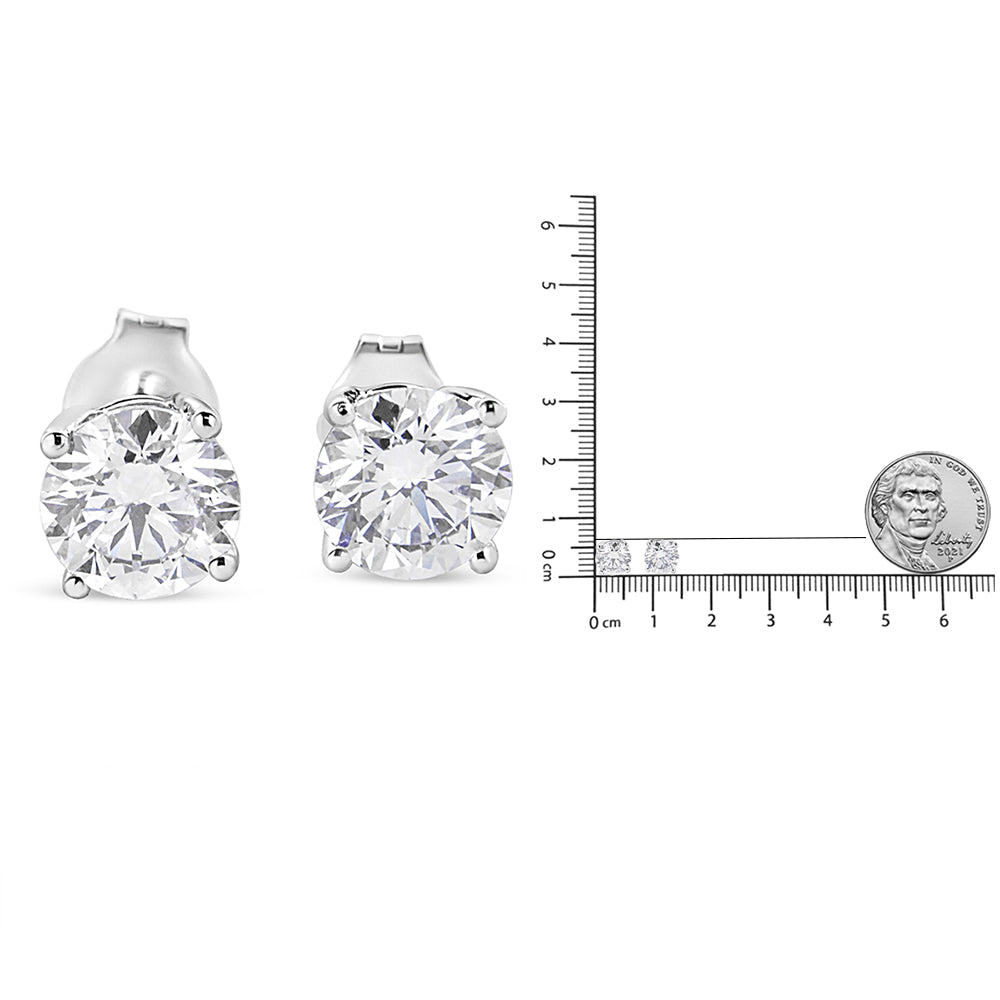 Front angle view of a pair of white metal stud earrings with round white gemstones in 4-prong settings, shown next to a ruler and coin for size reference.