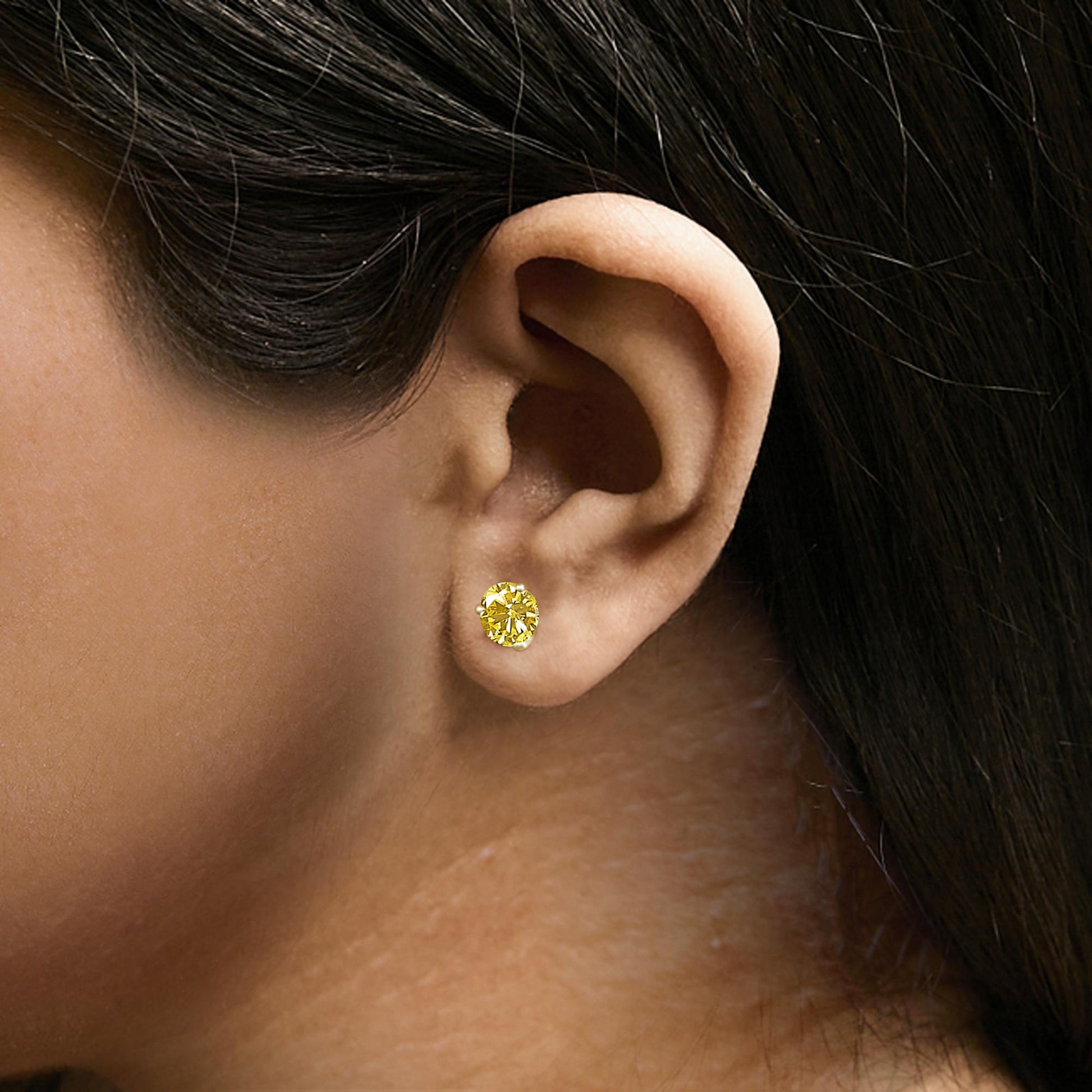 Close-up side view of a yellow metal stud earring with a round yellow gemstone in a 3-prong setting worn by a model.