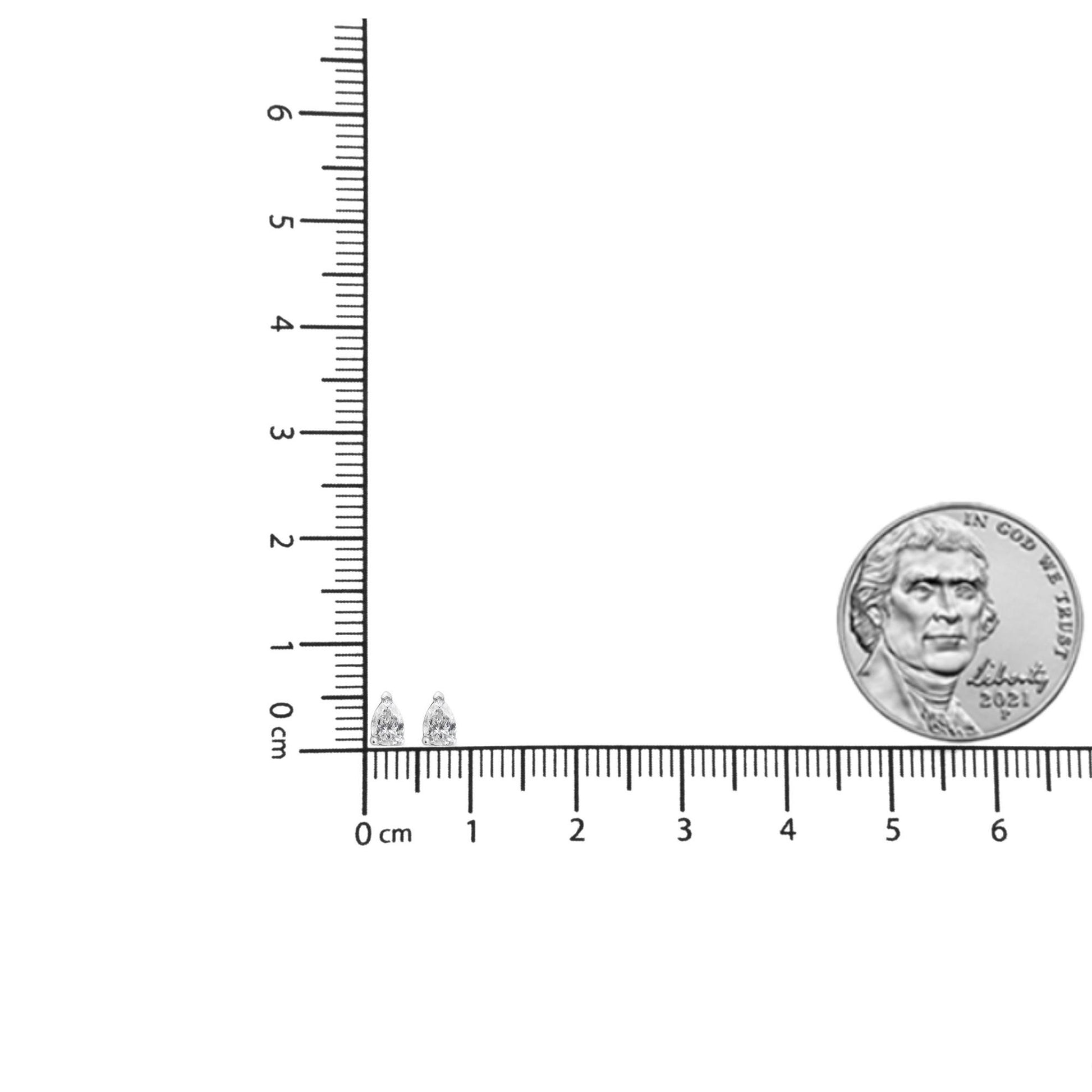 Pair of white metal pear-shaped earrings with white gemstones in 3-prong setting shown on a ruler and next to a coin for size reference, front angle.