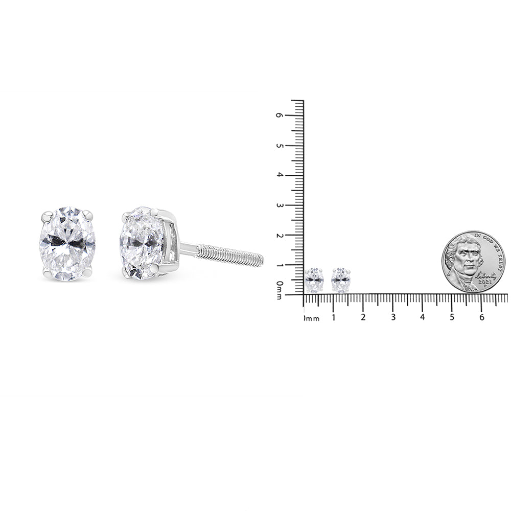Close-up front and side view of white metal stud earrings with oval-shaped white gemstones set in four-prong settings, displayed next to a ruler and a coin for size reference.