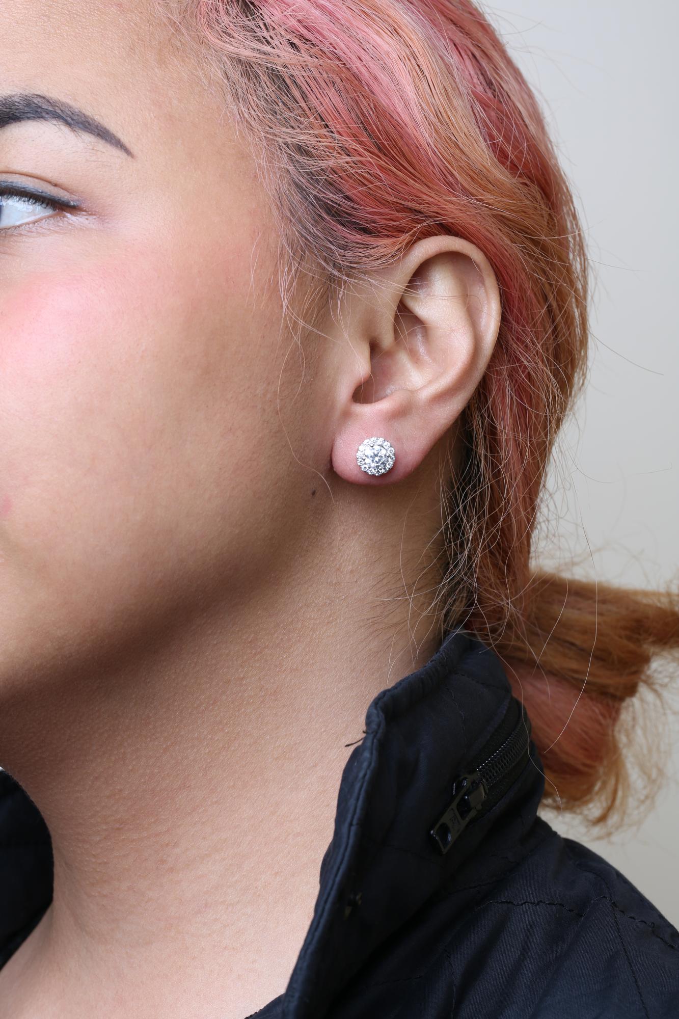 Close-up side view of white metal stud earrings with a central round white gemstone surrounded by smaller round white gemstones, worn on a model.