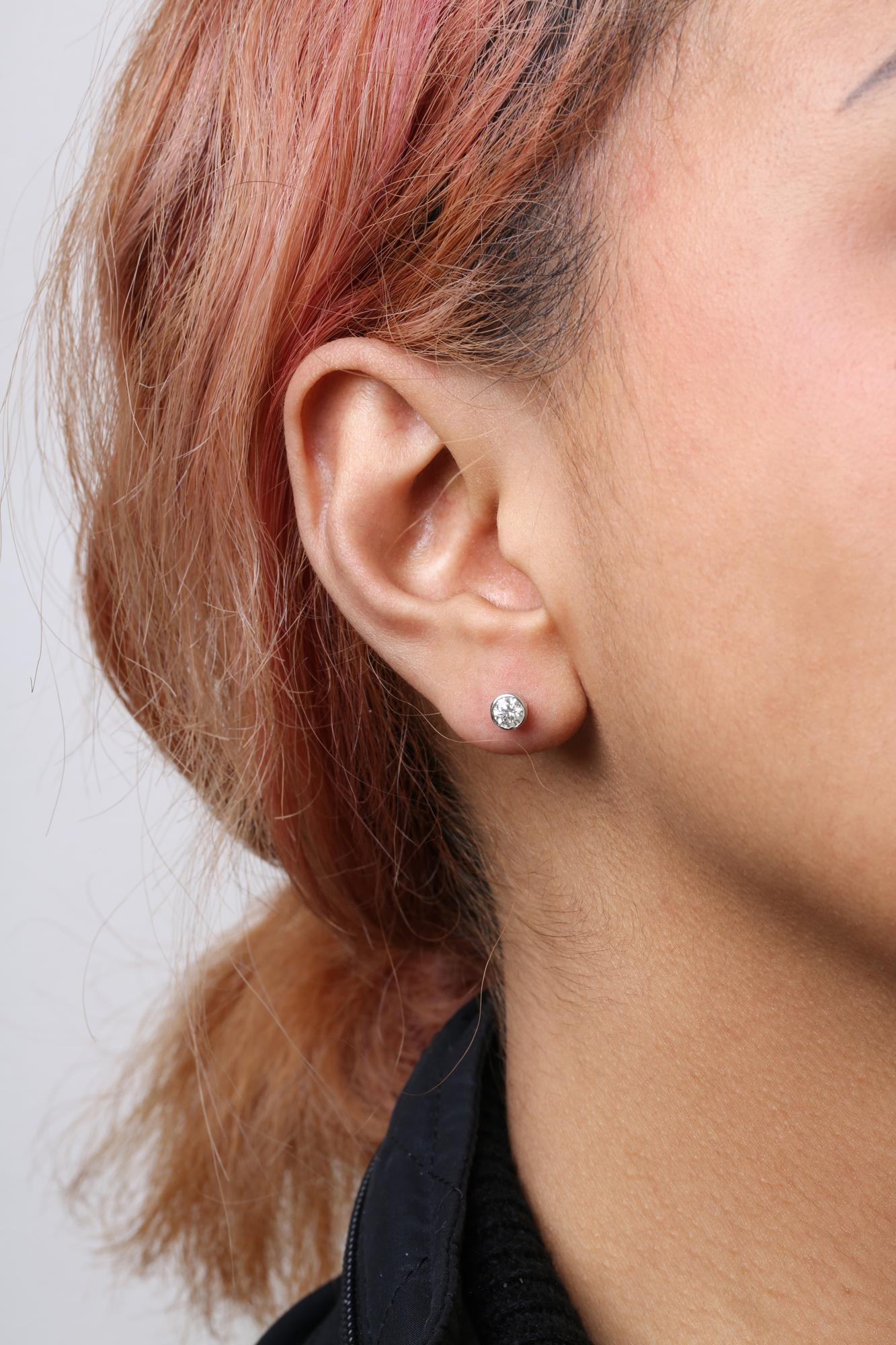 Close-up side view of an ear on a model wearing a white metal stud earring with a round white gemstone in a bezel setting.