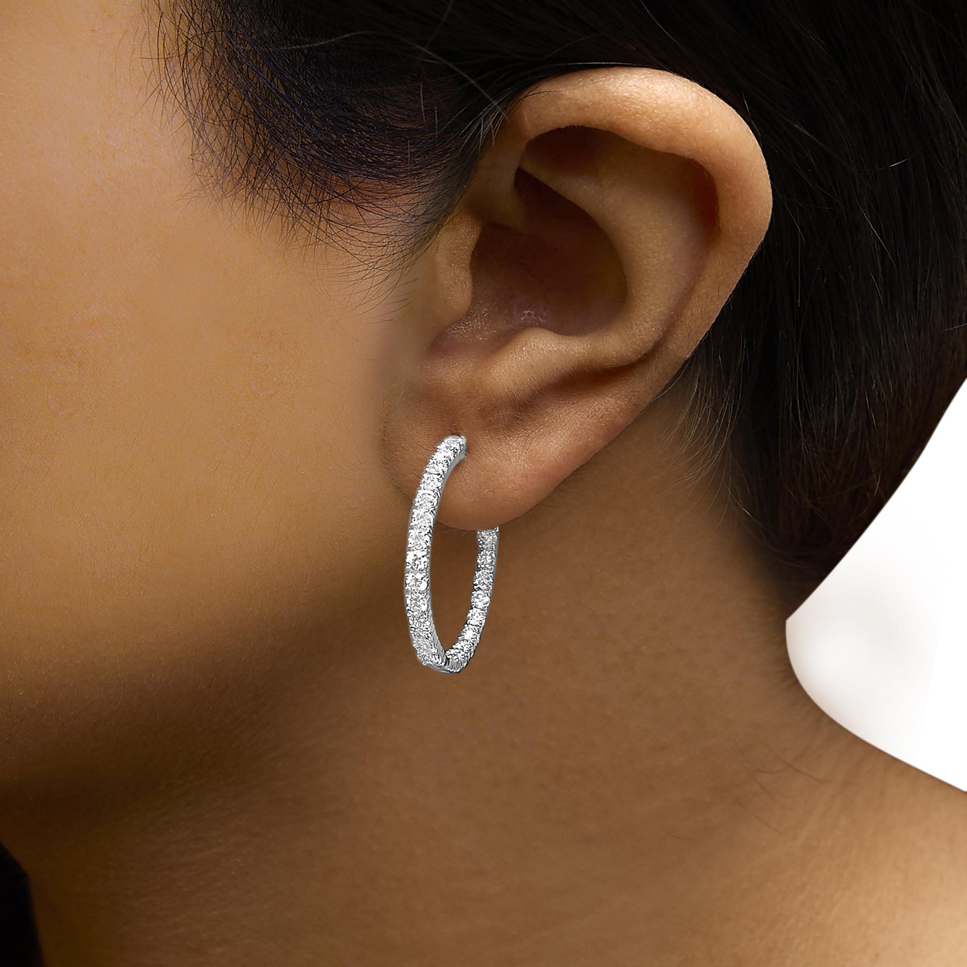 Close-up side view of a white metal hoop earring encrusted with round white gemstones, worn on a model's ear.