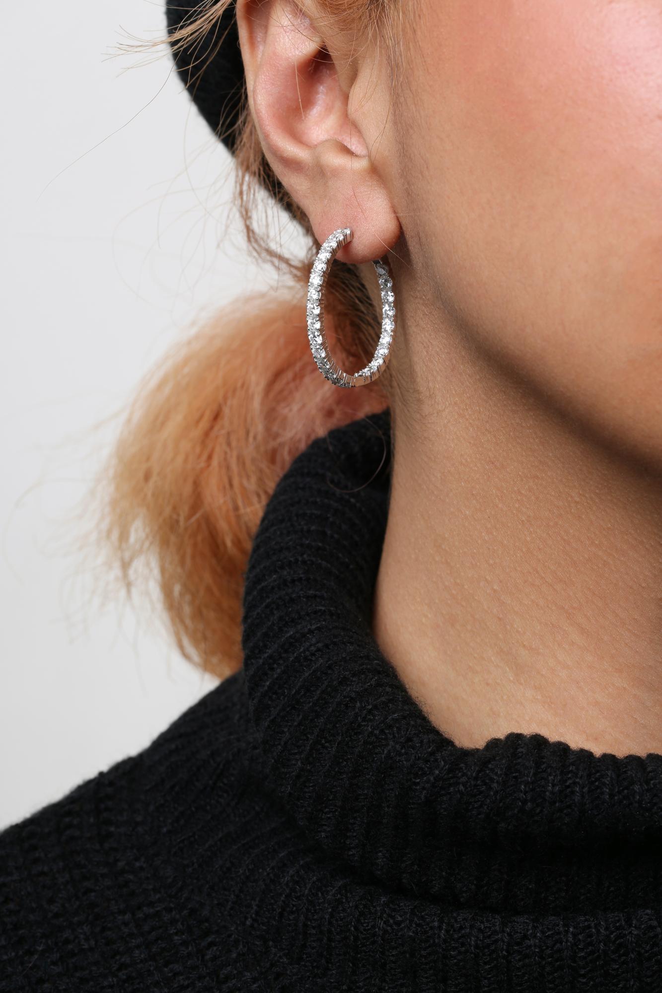 Close-up side view of a white metal hoop earring with round white gemstones in prong settings, worn on a model's ear.