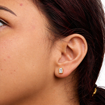 Close-up side view of a model wearing a yellow metal earring with an emerald-shaped white gemstone in a four-prong setting.