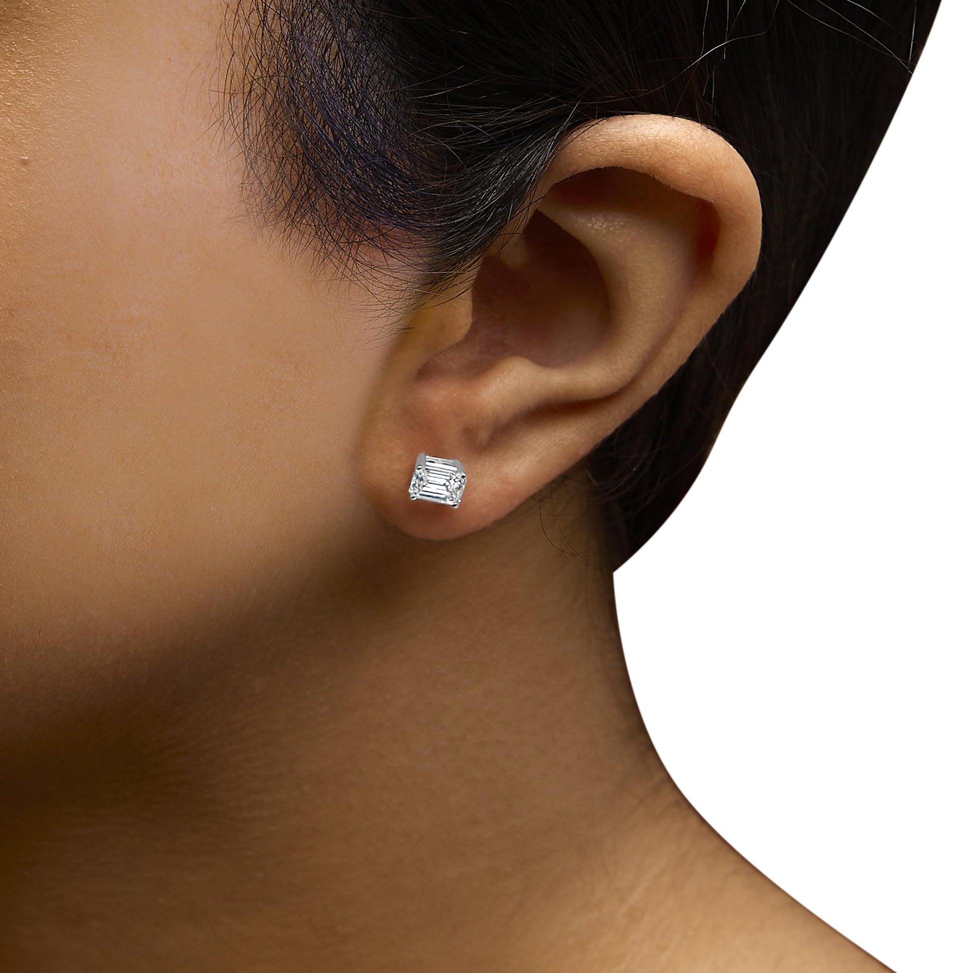 Close-up side view of a white metal earring with a single emerald-shaped white gemstone in a four-prong setting, worn on a model.