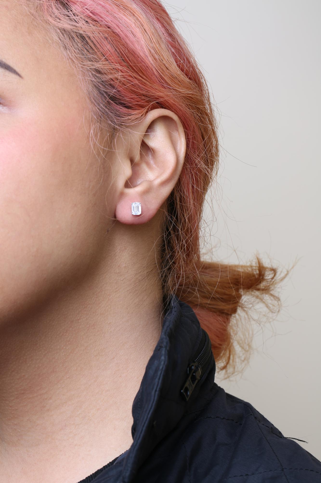 Close-up side view of a white metal earring with an emerald-shaped white gemstone in a four-prong setting, worn on a model.