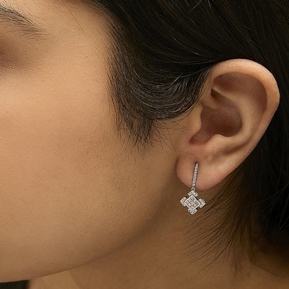 Close-up side view of a model wearing white metal earrings featuring clusters of princess-shaped and round white gemstones, with the earrings hanging below the earlobe.