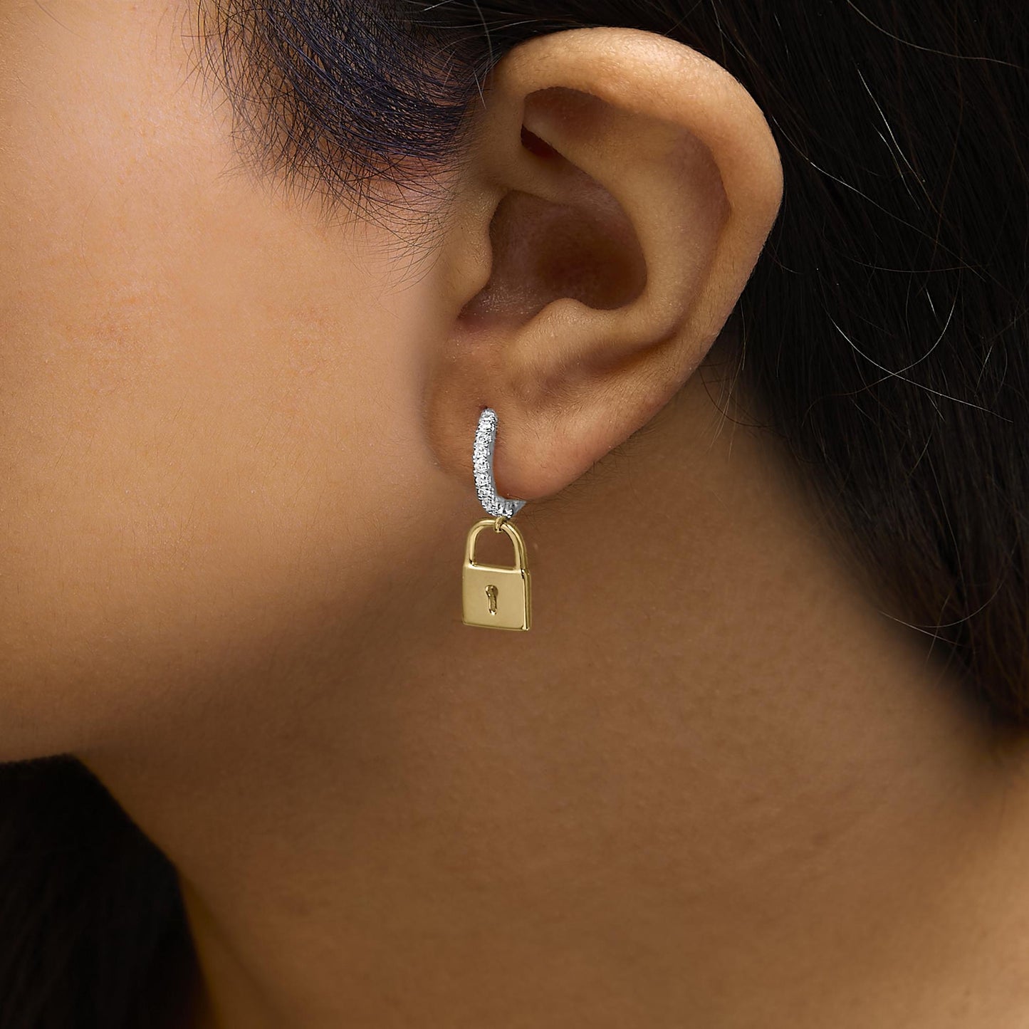 Close-up side view of a yellow metal earring on a model, featuring a curved section with white round gemstones in prong settings and a small padlock-shaped charm hanging below.