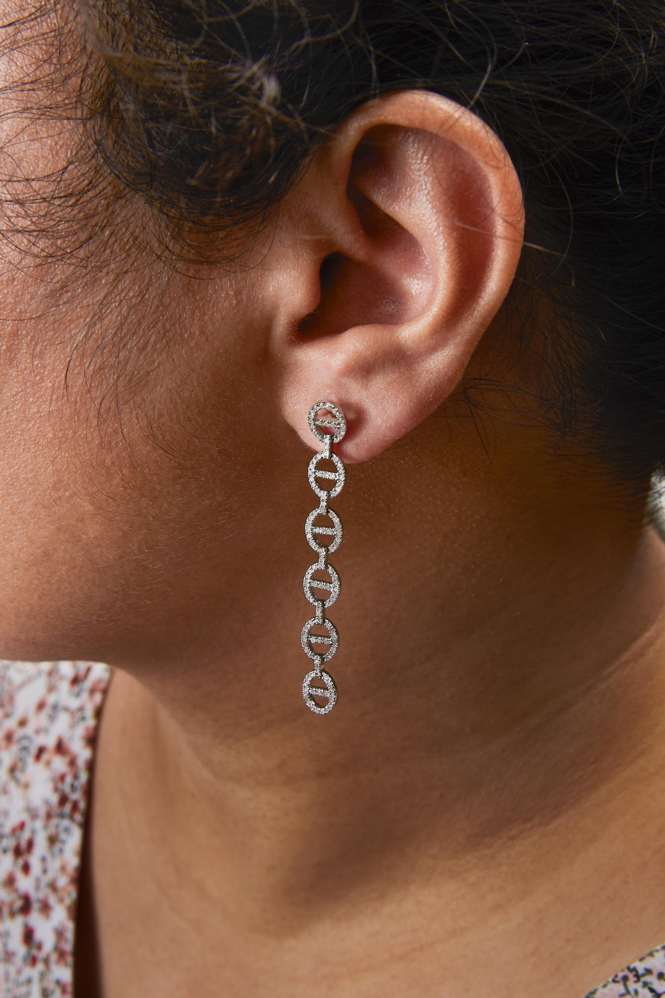 Close-up side view of a model wearing long white metal earrings featuring linked circular shapes encrusted with small round white gemstones in prong settings.