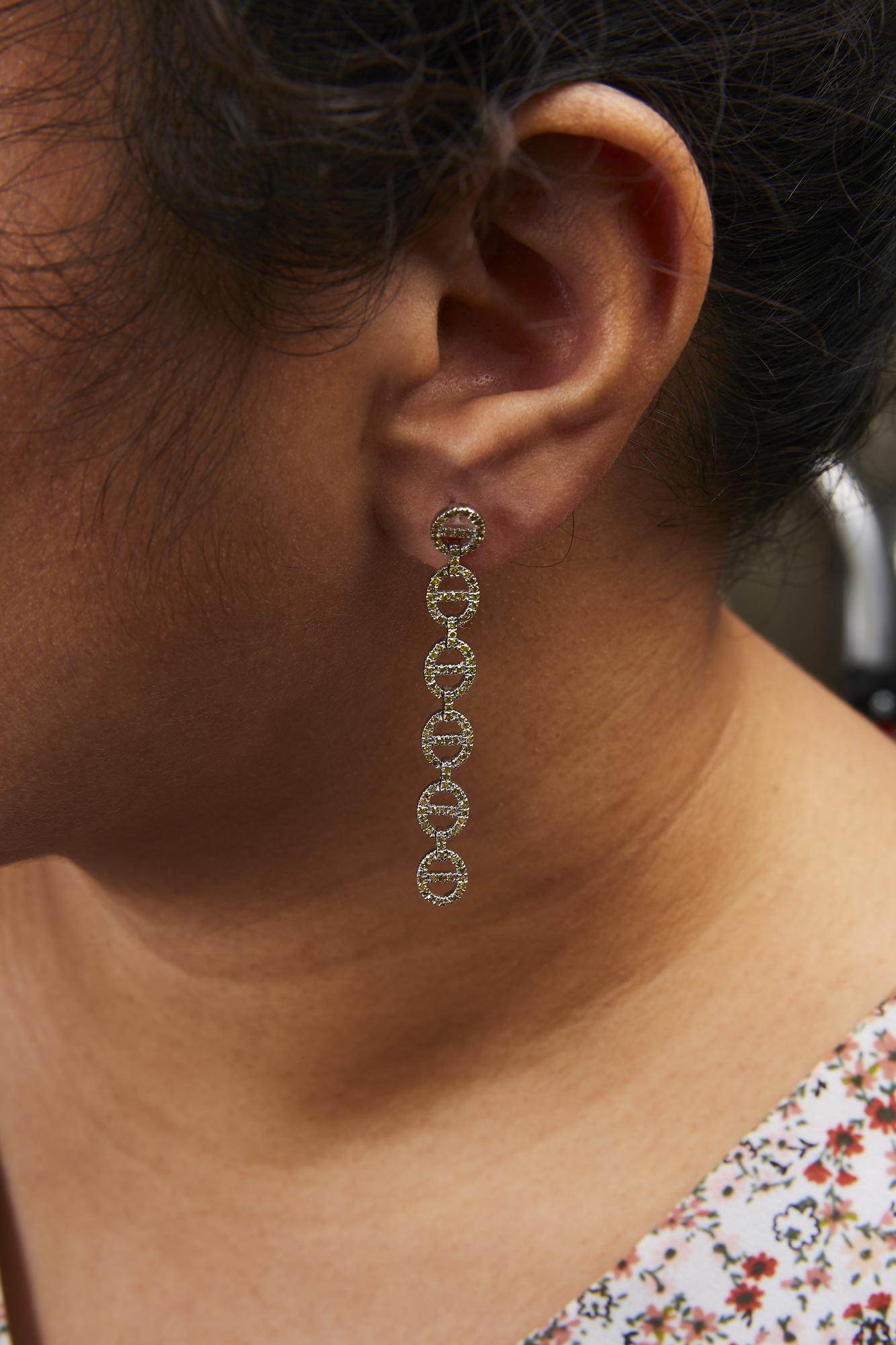 Close-up side view of a model wearing a long white metal earring composed of linked circular shapes, each adorned with small yellow round gemstones in prong settings.