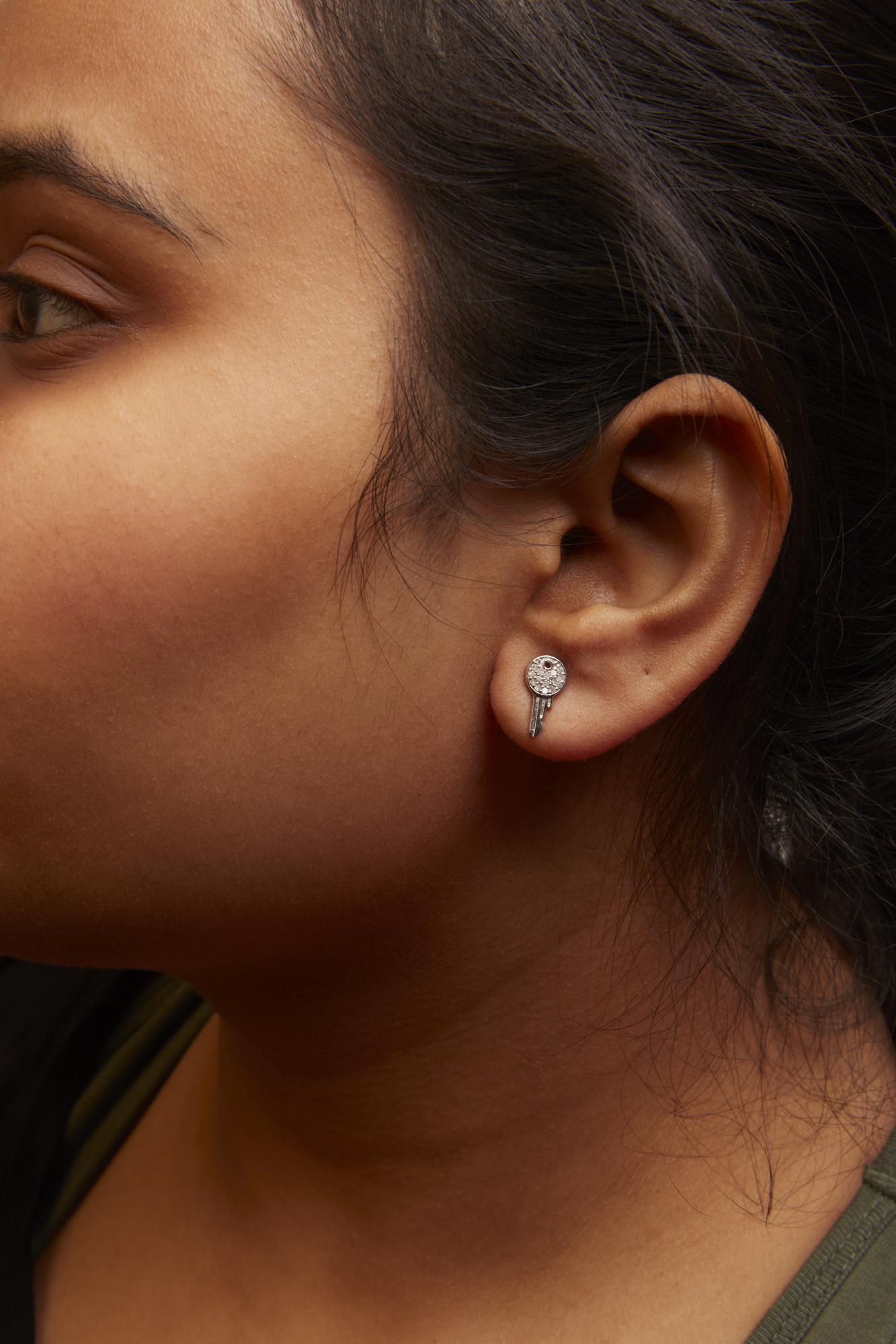Close-up side view of a model wearing white metal earrings with a circular top encrusted with white gemstones and a small key-shaped dangle.