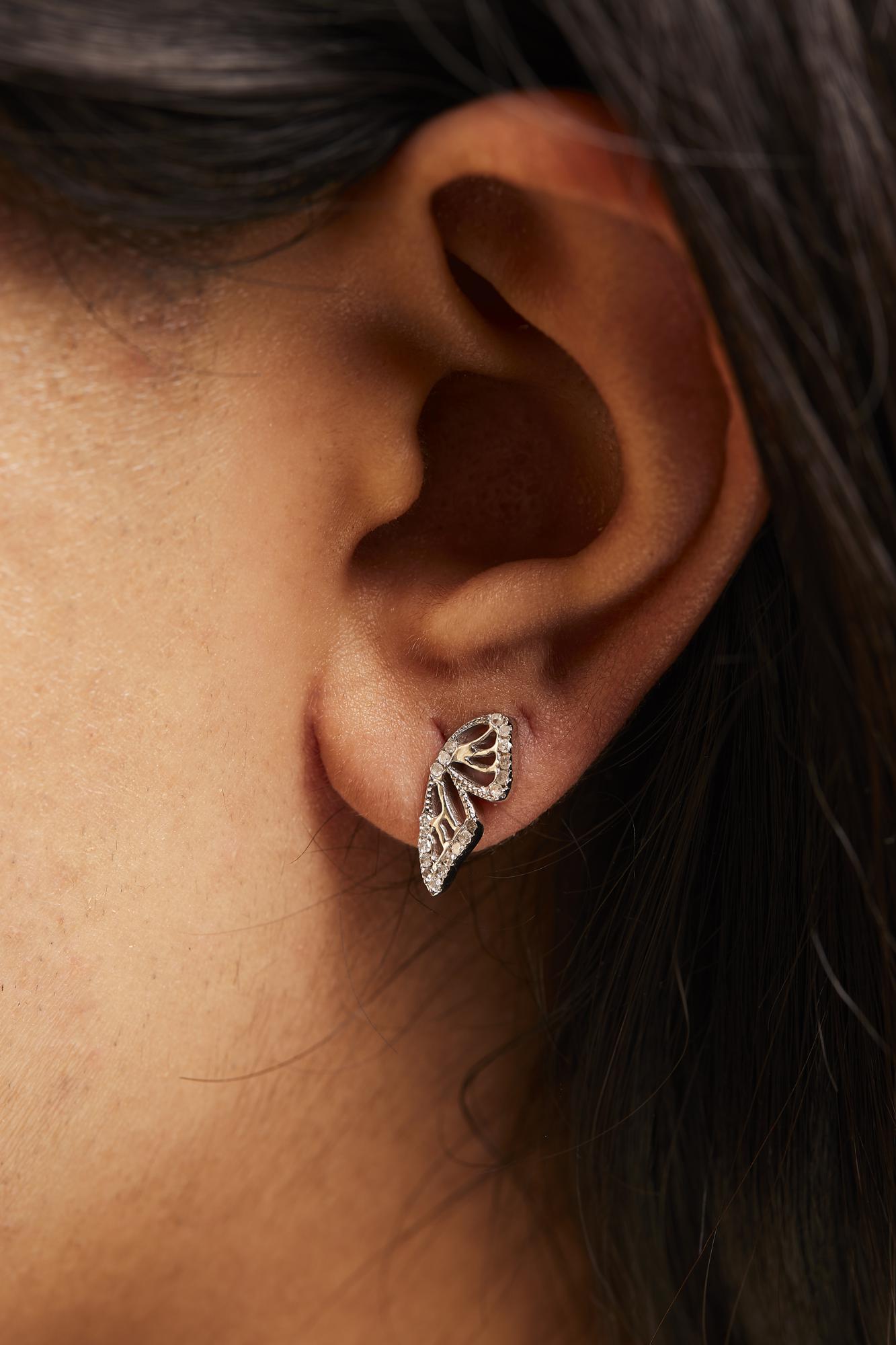 Close-up side view of a white metal earring shaped like two connected abstract leaves with round white gemstones in prong settings, shown worn on a model's ear.