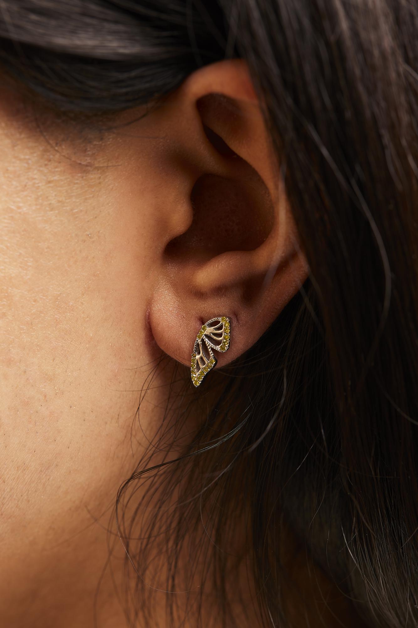 Close-up side view of a model wearing white metal earrings shaped like stylized leaves with yellow round prong-set gemstones outlining the edges.
