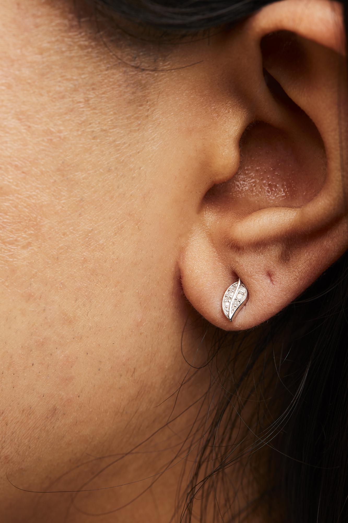 Close-up side view of a model wearing white metal earrings shaped like leaves, featuring multiple small round white gemstones in prong settings.