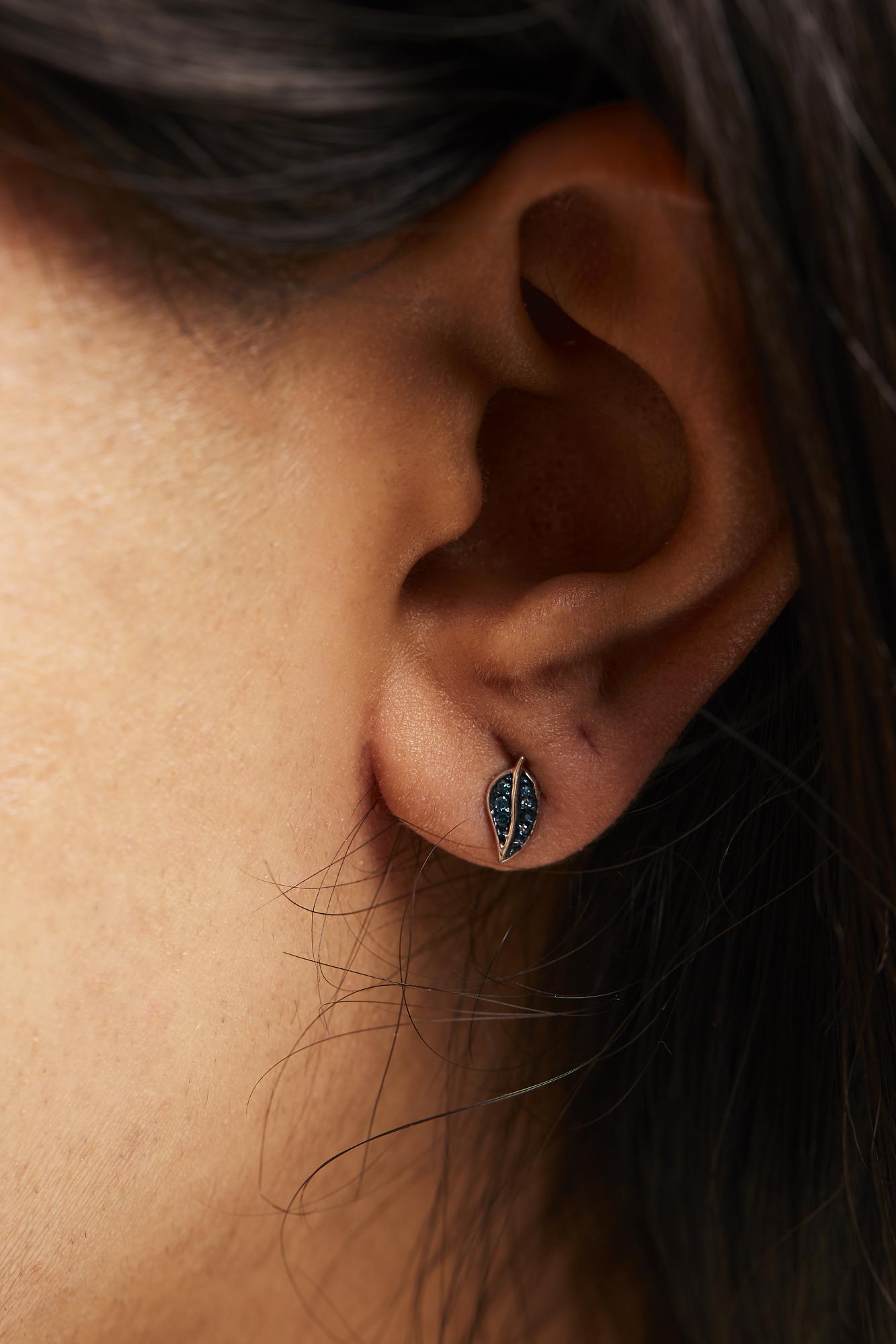 Close-up side view of a white metal earring worn on a model's ear, featuring an elongated almond shape with multiple blue round prong-set gemstones clustered inside.