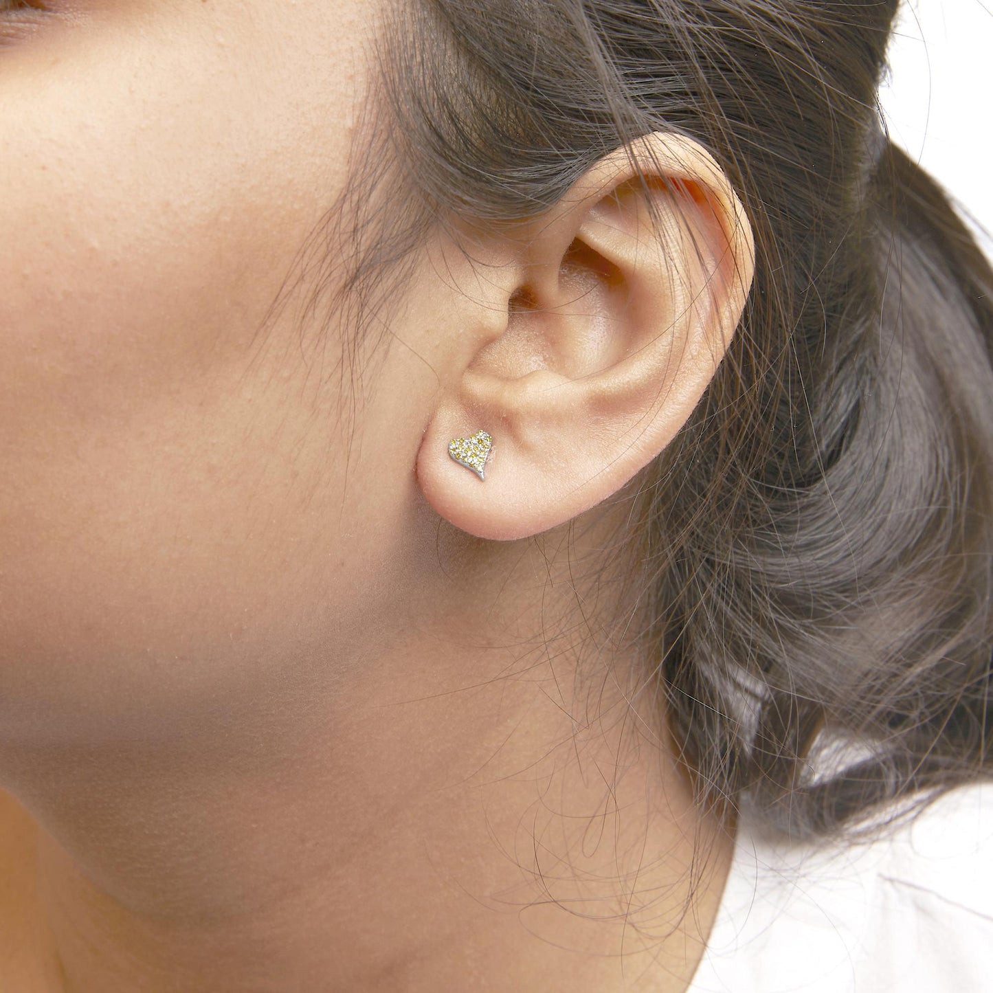 Close-up side view of a model wearing white metal heart-shaped stud earrings with yellow round gemstones in prong settings.