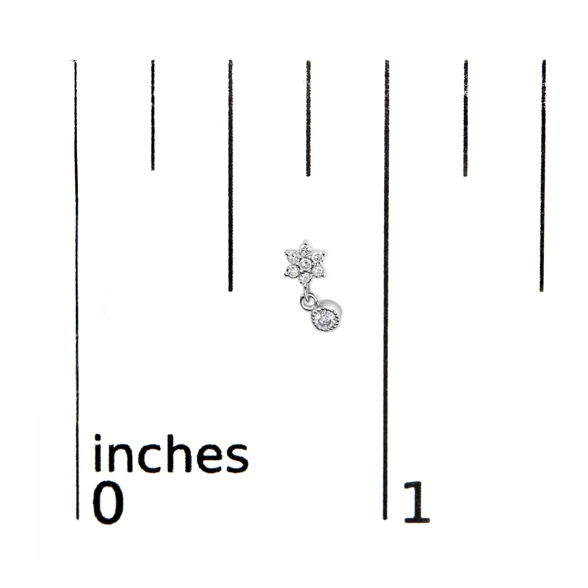 Close-up of white metal earring with small round prong-set white gemstones arranged in a flower shape and a single gemstone dangle, placed on a ruler for size reference.