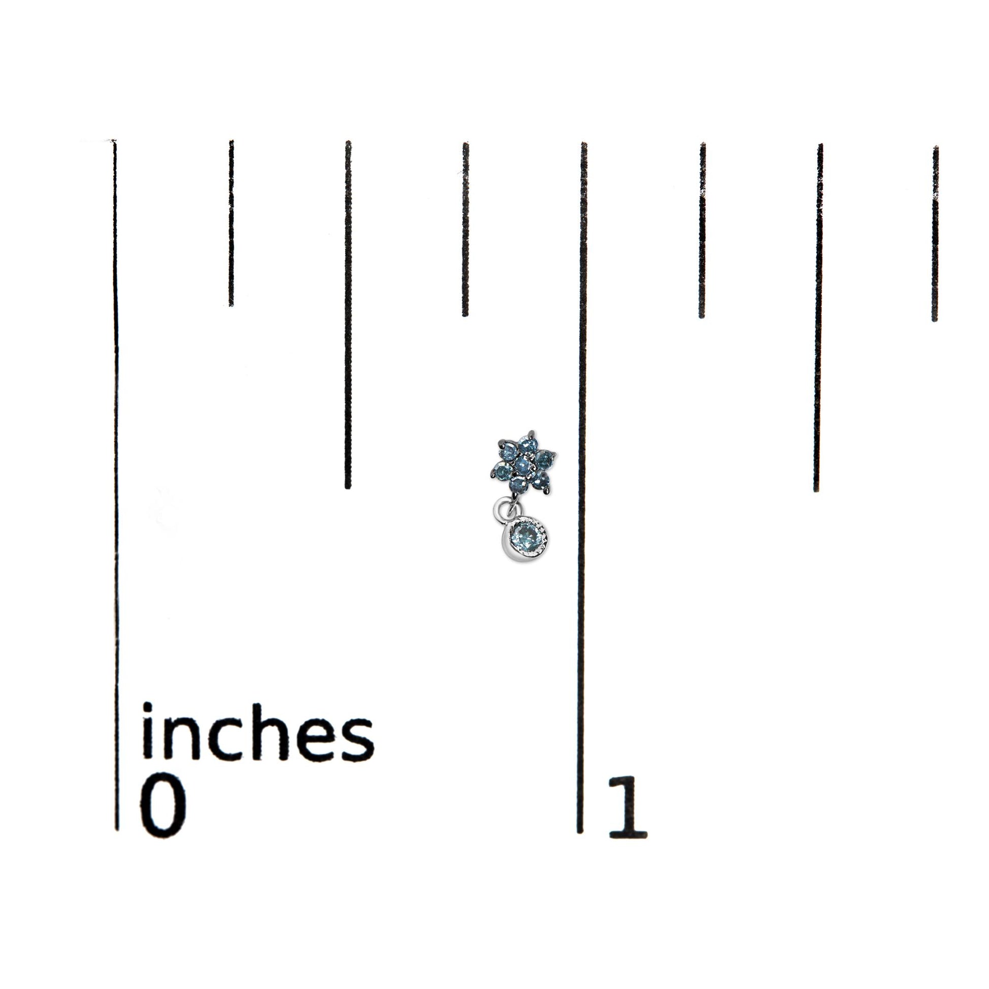White metal earrings with blue round prong-set gemstones, shaped like a small flower with a dangling gemstone, shown on a ruler for size reference, top-down angle.