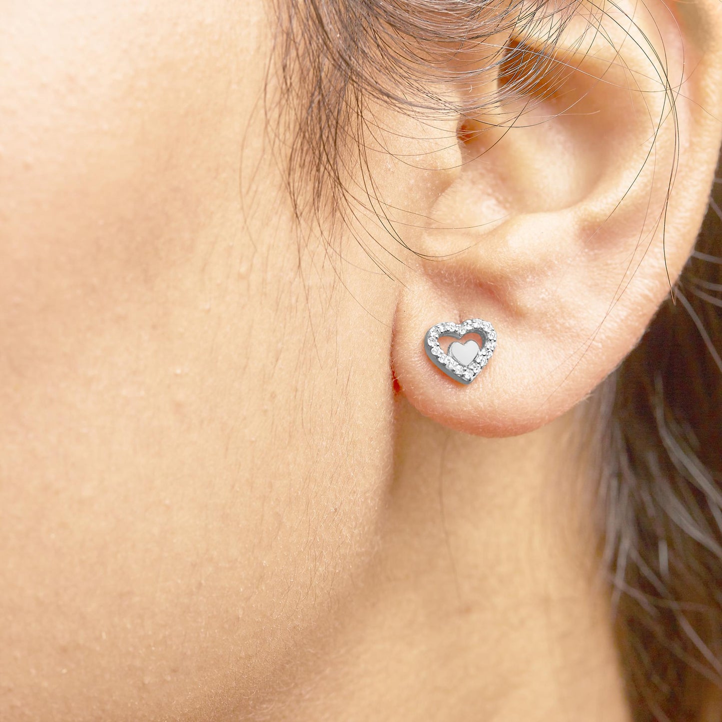 Close-up side angle of a white metal heart-shaped earring with round prong-set white gemstones worn on a model's ear.