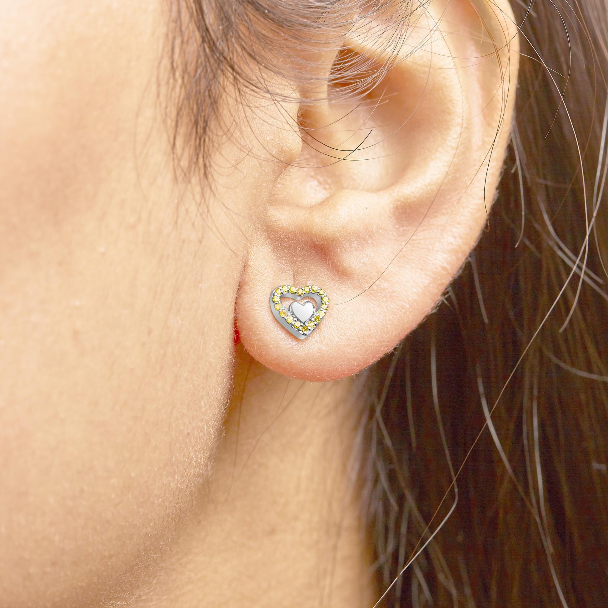 Close-up side view of a white metal heart-shaped stud earring with a border of yellow round prong-set gemstones, worn on a model's ear.