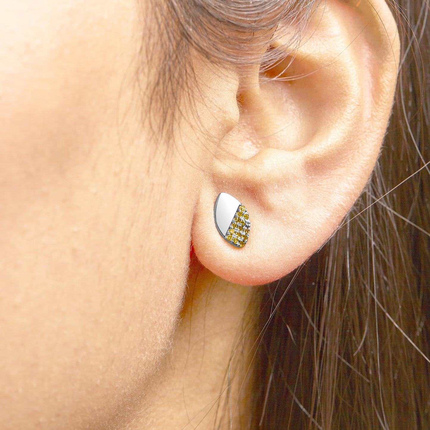 Close-up side view of a white metal earring on a model's ear, featuring a half smooth surface and half covered with small yellow gemstones in a prong setting.