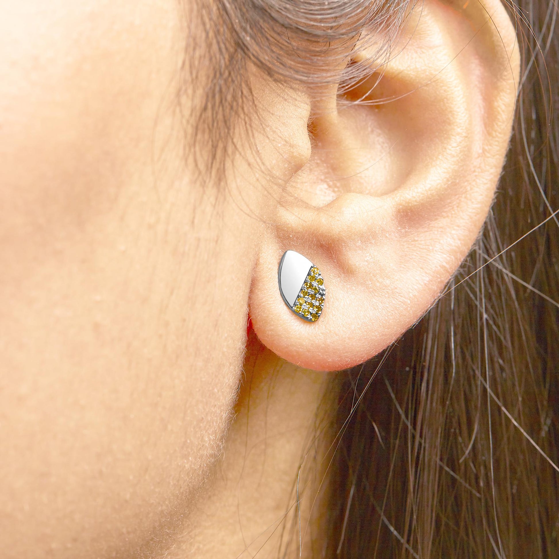 Close-up side view of a white metal earring on a model's ear, featuring a half smooth surface and half covered with small yellow gemstones in a prong setting.