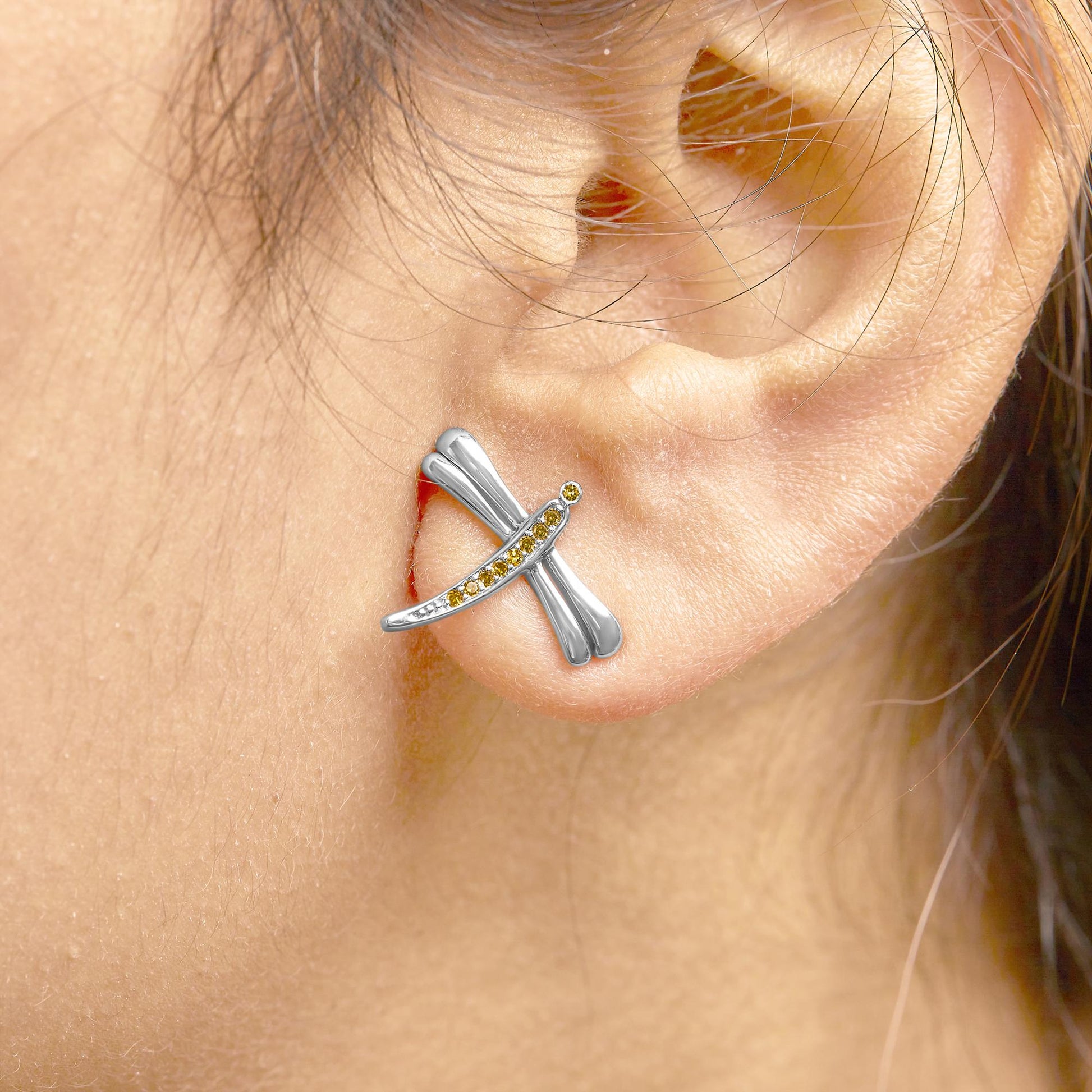 Close-up side view of white metal earrings on a model's ear, featuring crossing rounded bars with prong-set yellow gemstones.