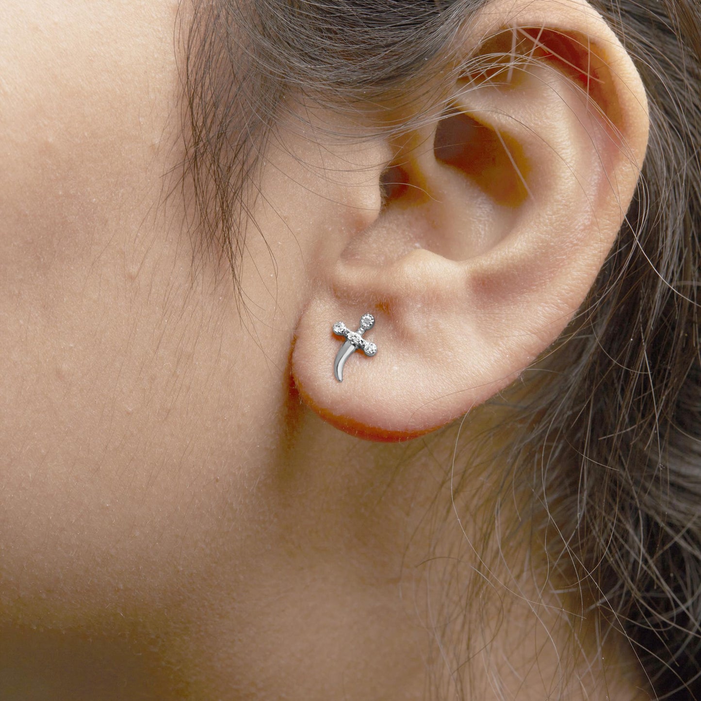 Close-up side view of a white metal earring on a model's ear, featuring a small cross shape with white round gemstones in prong settings.