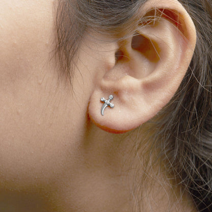 Close-up side view of a white metal earring on a model's ear, featuring a small cross shape with white round gemstones in prong settings.