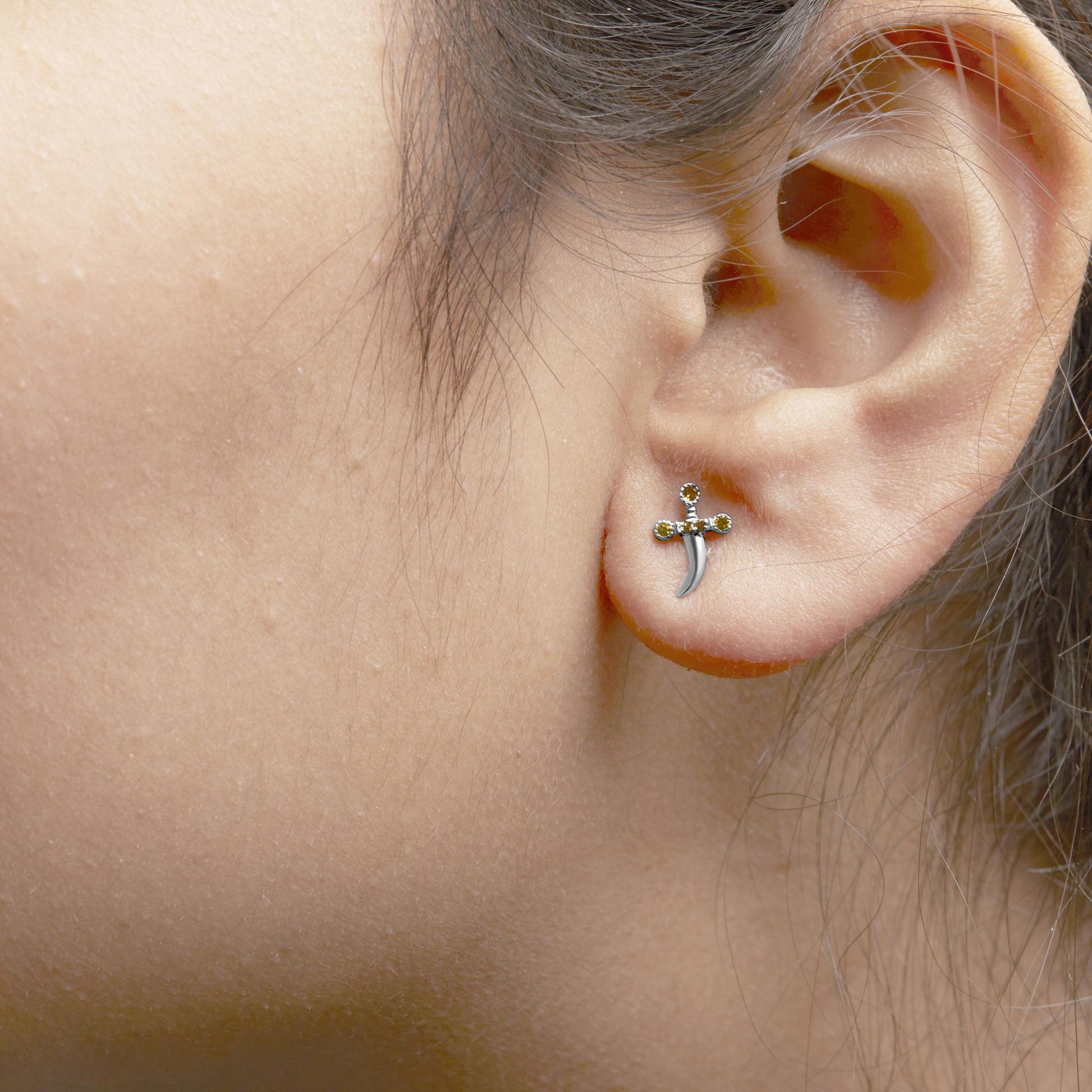 Close-up side view of a white metal earring on a model's ear, shaped like a small cross with round yellow gemstones in prong settings.