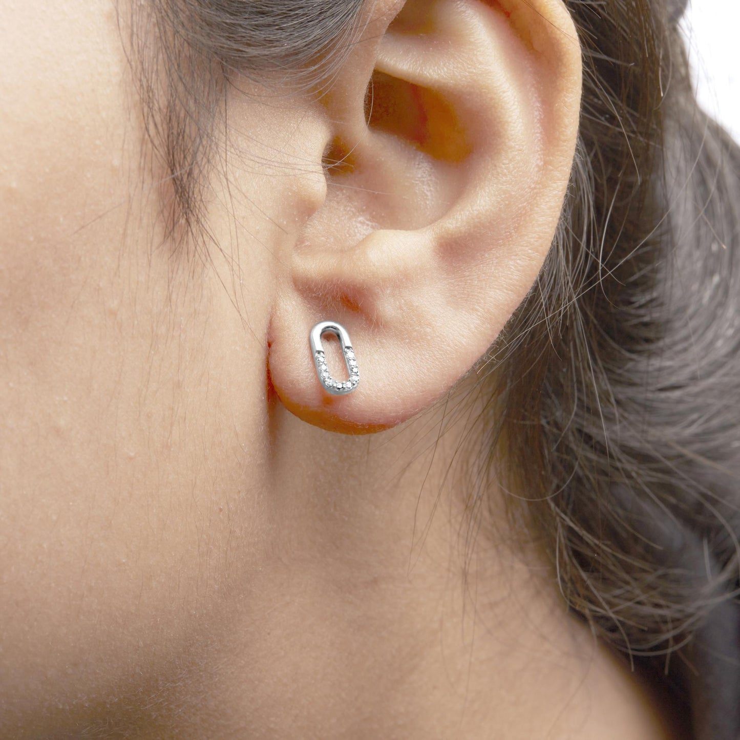 Close-up side view of a white metal earring on a model's ear featuring an elongated oval shape with round white gemstones set in prongs along the lower half.