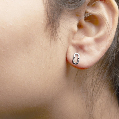 Close-up side view of a white metal earring on a model's ear, shaped like a vertical oval link with multiple small round blue gemstones set in the lower half.
