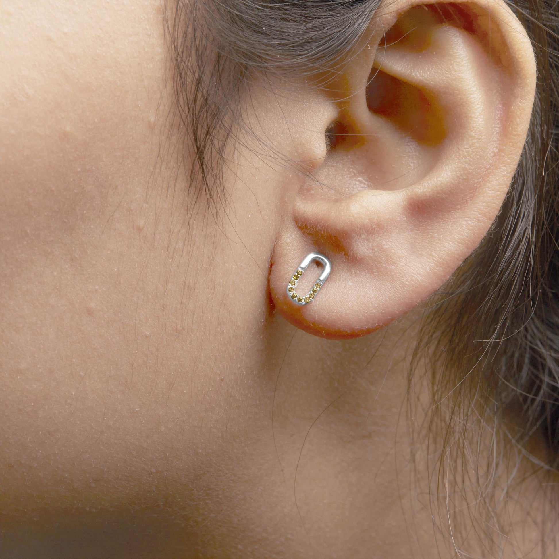 Close-up side view of an ear wearing a white metal oval-shaped earring adorned with yellow round gemstones in a prong setting along the bottom half.