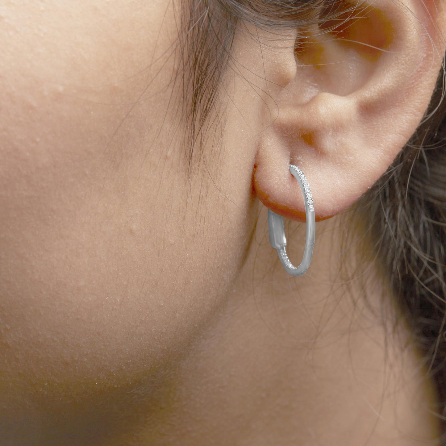 Close-up side view of a white metal hoop earring with round white gemstones in prong setting, worn on a model's ear.