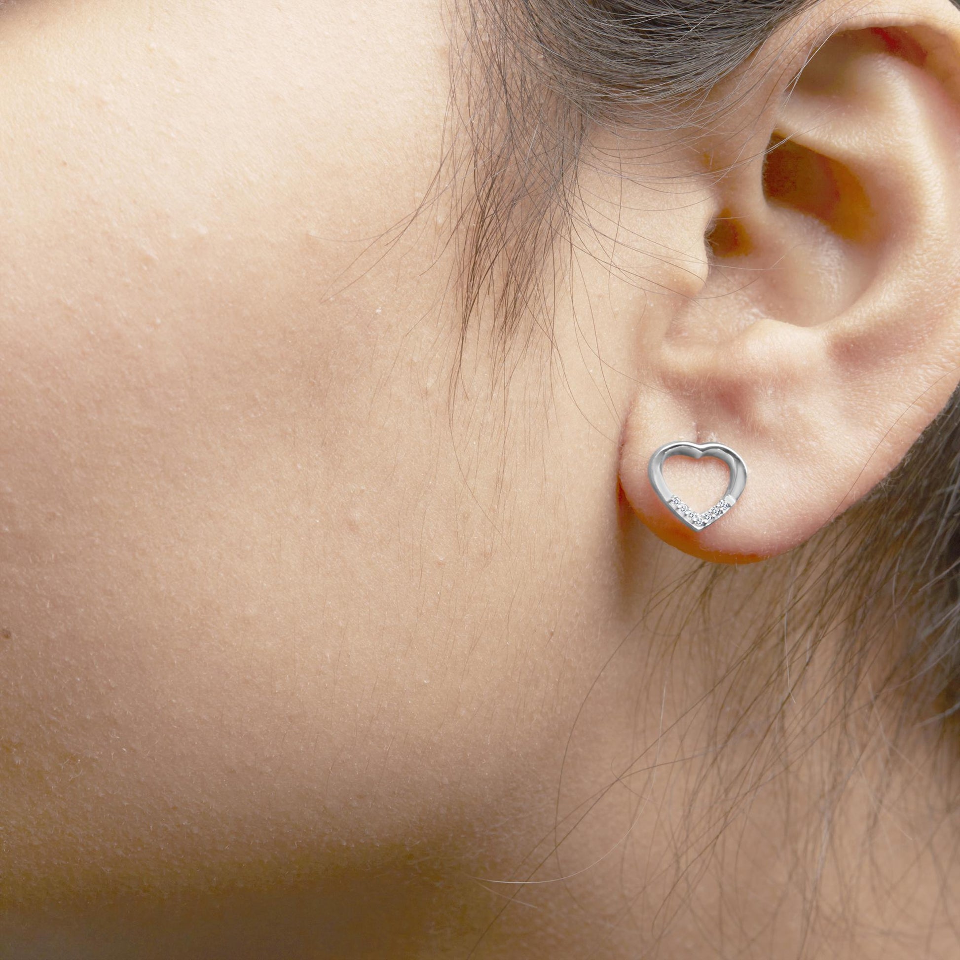 Close-up side view of a heart-shaped white metal earring with small round white gemstones in prong settings along the bottom curve, worn on a model's ear.