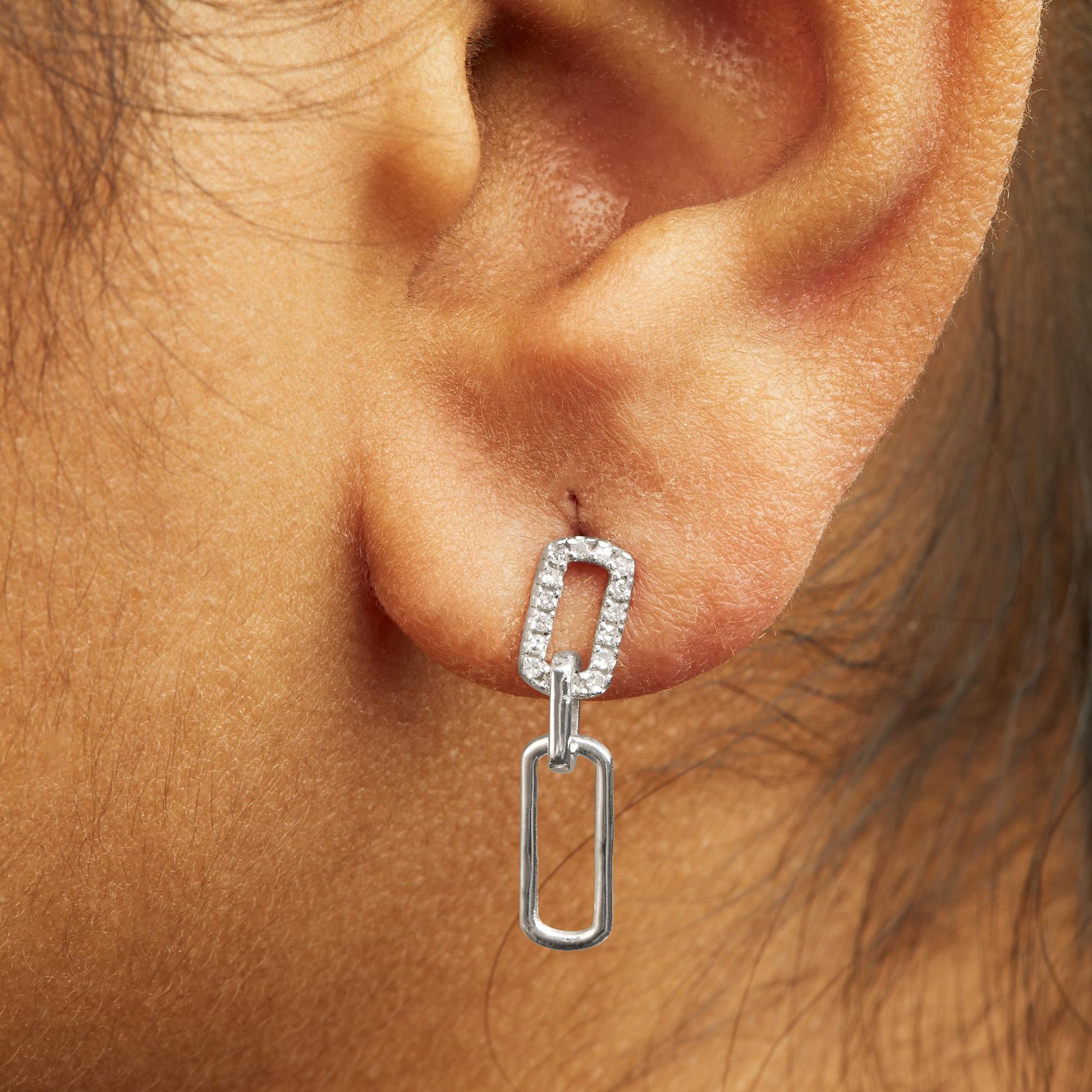 Close-up side view of a white metal earring on a model’s ear featuring a rectangular link with small round white gemstones in prong settings connected to a larger open rectangular link.