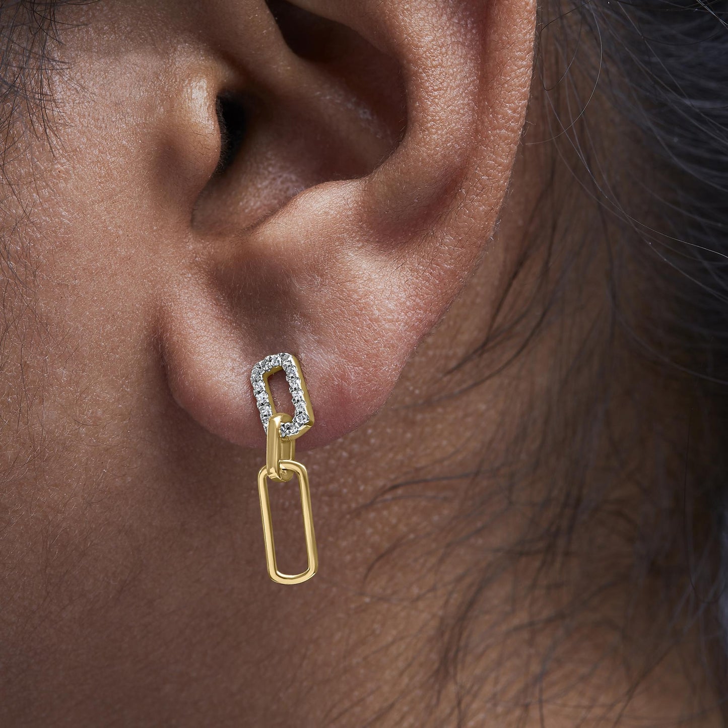 Close-up side view of a model wearing yellow metal earrings with two linked rectangular shapes, the top rectangle adorned with white round prong-set gemstones.