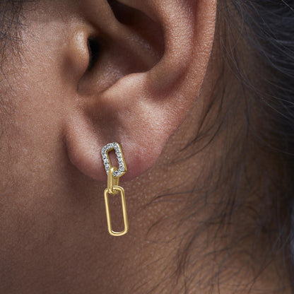 Close-up side view of a model wearing yellow metal earrings with two linked rectangular shapes, the top rectangle adorned with white round prong-set gemstones.