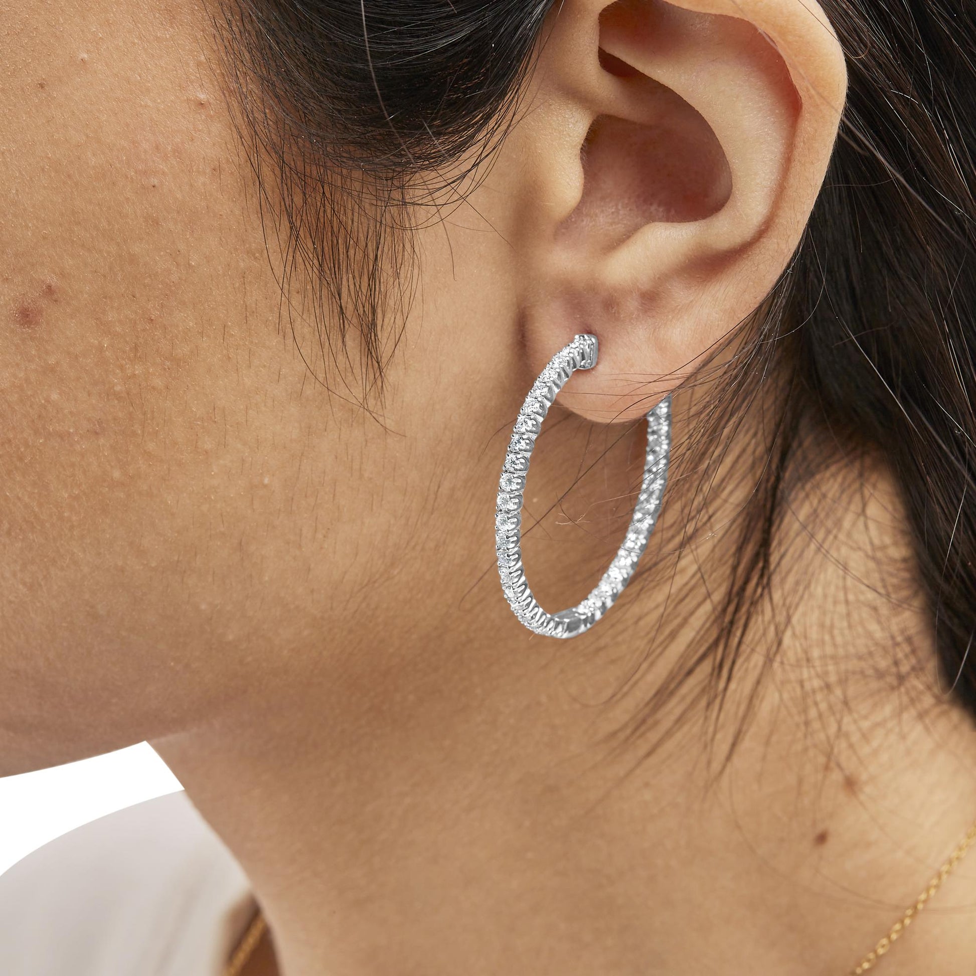 Close-up side view of a white metal hoop earring adorned with round white gemstones in prong settings, worn on a model.