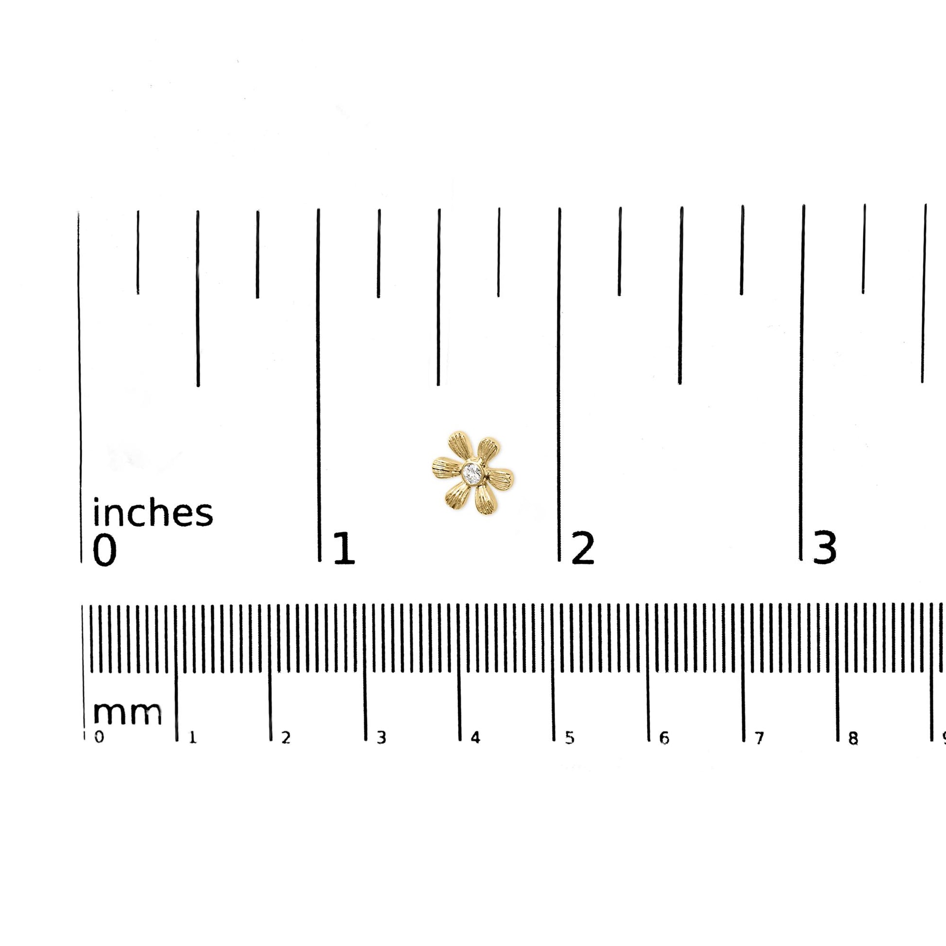 Close-up of a yellow metal flower-shaped earring with a round white gemstone in the center, placed on a ruler for size reference.
