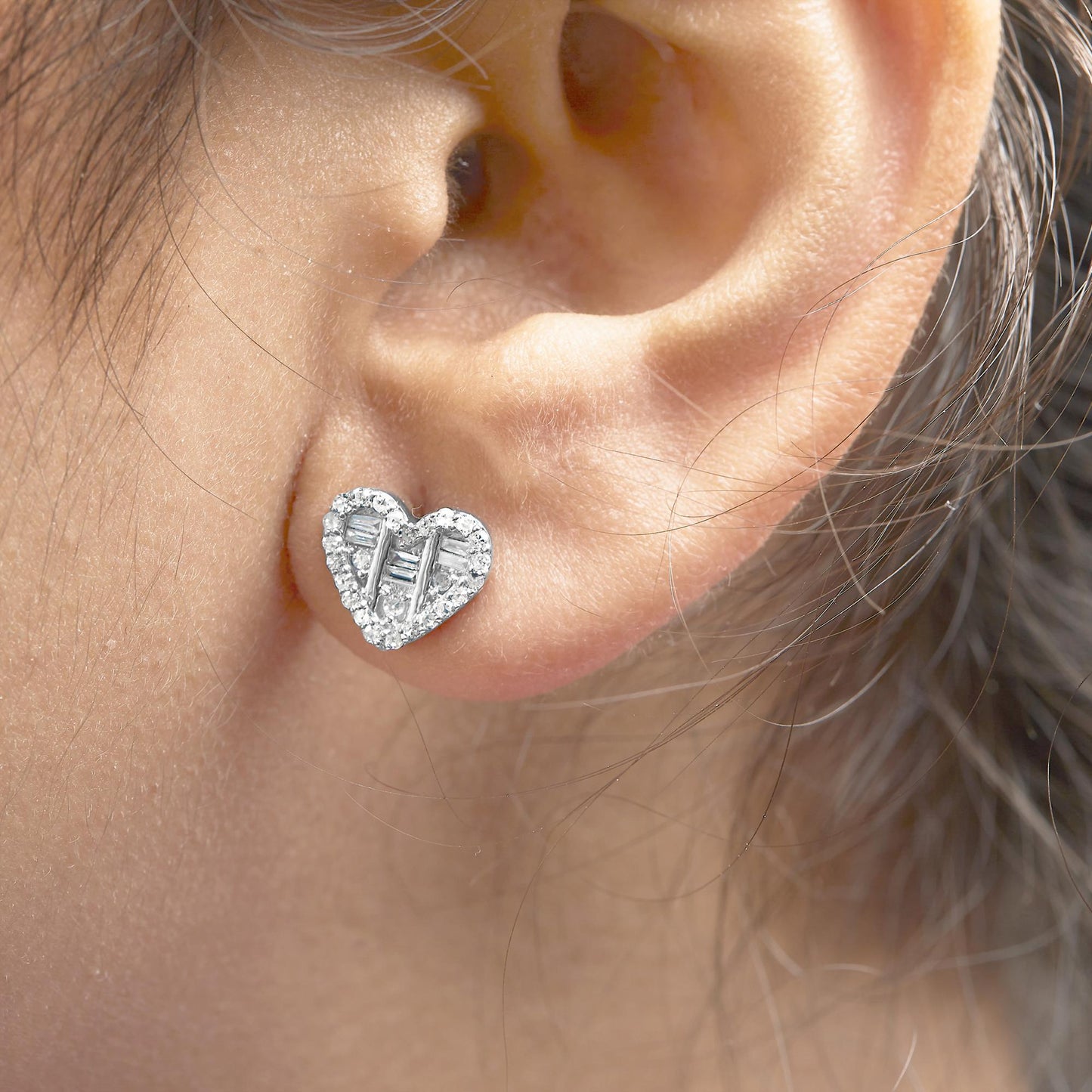 Close-up side angle of a white heart-shaped earring on a model's ear, featuring round and baguette-shaped white gemstones in prong and channel settings.