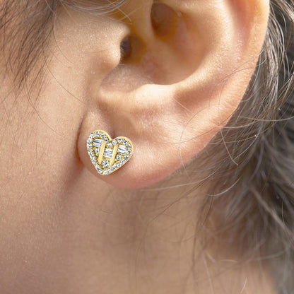 Close-up side view of a heart-shaped yellow metal earring on a model's ear, featuring small round white gemstones in prong settings outlining the heart shape, and three vertical rows of baguette-shaped white gemstones in channel settings inside the heart.