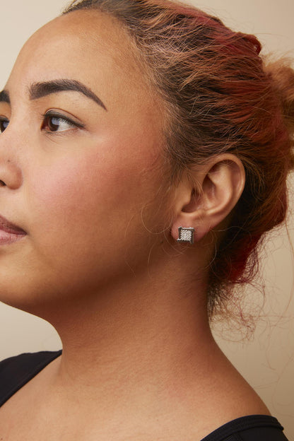 White metal square-shaped stud earrings with prong-set white gemstones, worn on a model shown in a side profile view.