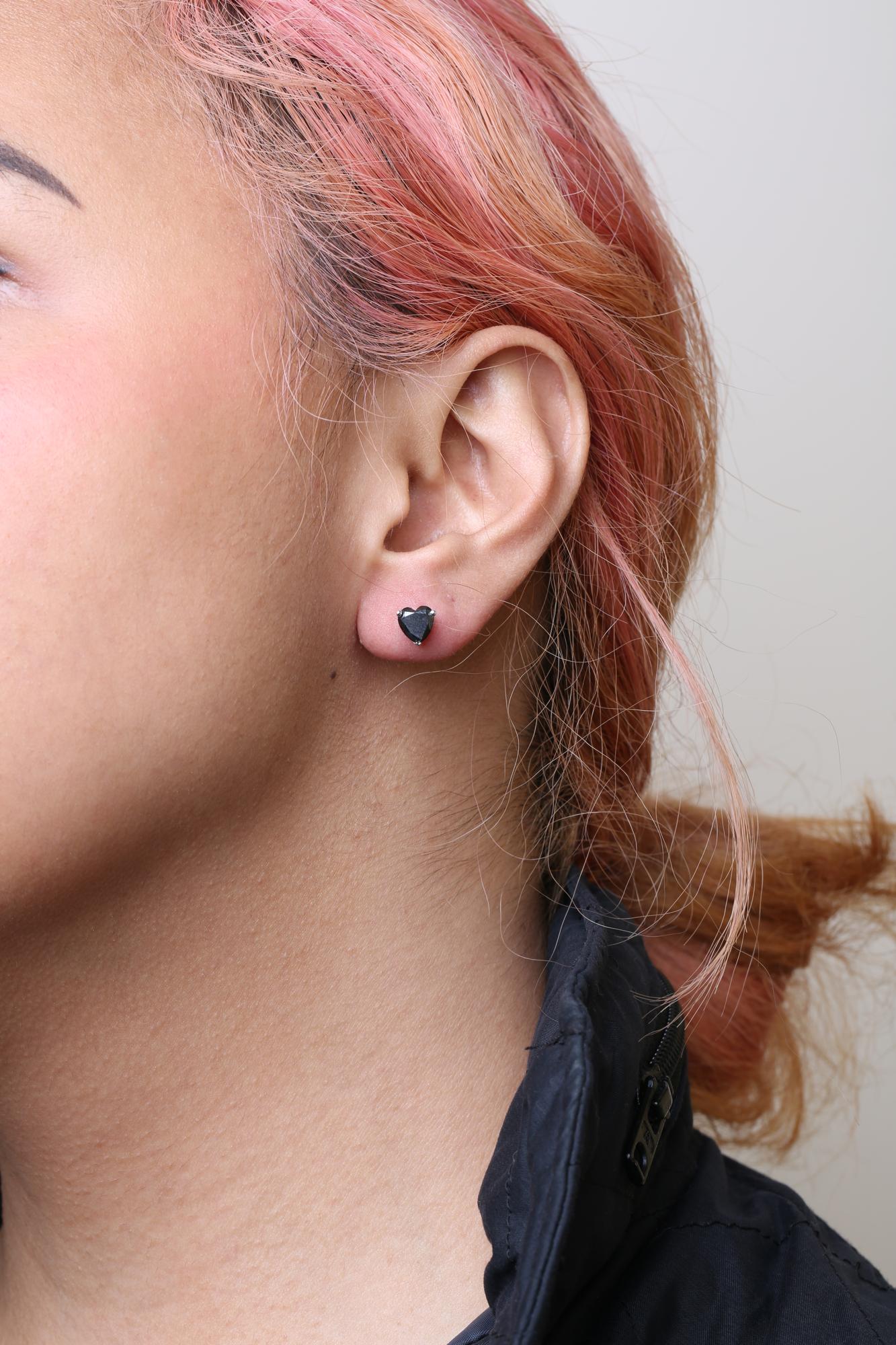 Close-up side view of a model wearing a white metal stud earring with a heart-shaped black gemstone in a prong setting.