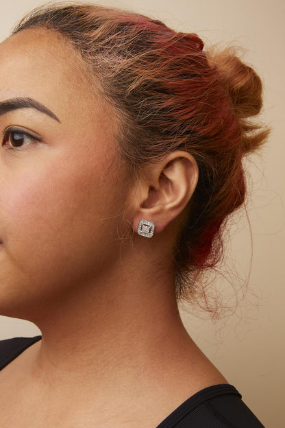 Close-up side view of a model wearing square-shaped white metal earrings featuring white princess-shaped gemstones in an invisible setting surrounded by round white gemstones in prong settings.