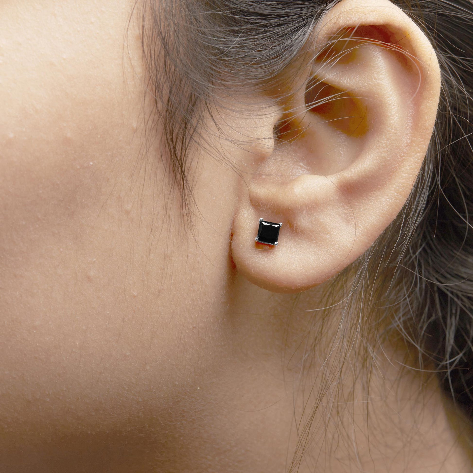Close-up side view of a white metal earring with a black princess-shaped gemstone in a 4-prong setting worn on a model's ear.